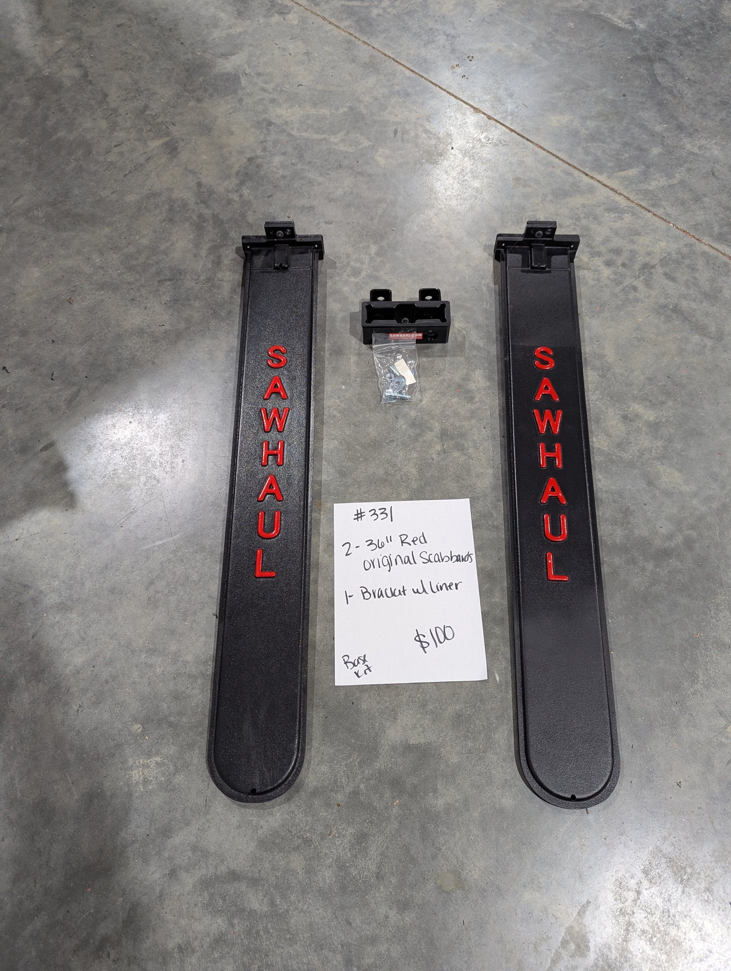 Two black 'SawHaul' 36 inch scabbards and a bracket with liner on a gray floor with a price tag. On closeout Lot 2