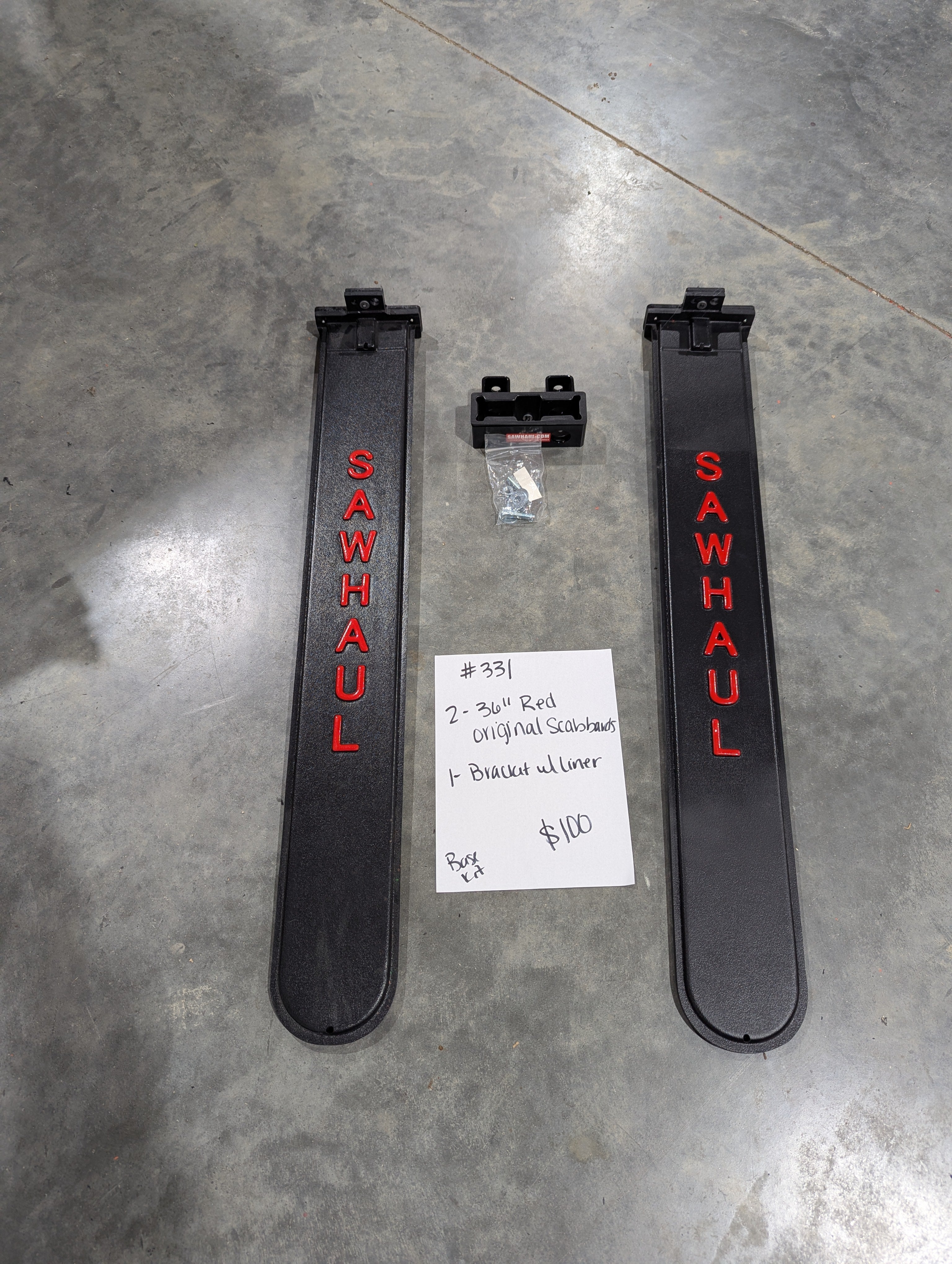 Two black 'SawHaul' 36 inch scabbards and a bracket with liner on a gray floor with a price tag. On closeout Lot 2